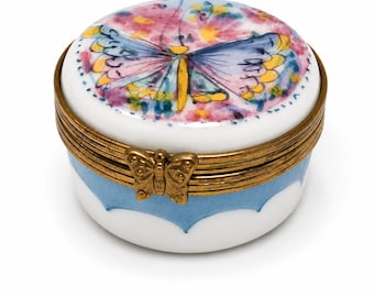 May include: A round white porcelain trinket box featuring a hand-painted butterfly with blue, pink, and yellow wings on the lid. The box has a brass metal hinge and rim, with a small butterfly-shaped clasp closure and a light blue scalloped base.