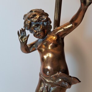 May include: A bronze-toned metal lamp base featuring a sculpted figure of a cherub with curly hair, arms raised in a dynamic pose, and a draped cloth around the waist. The figure is attached to a vertical fluted metal rod with a light socket.