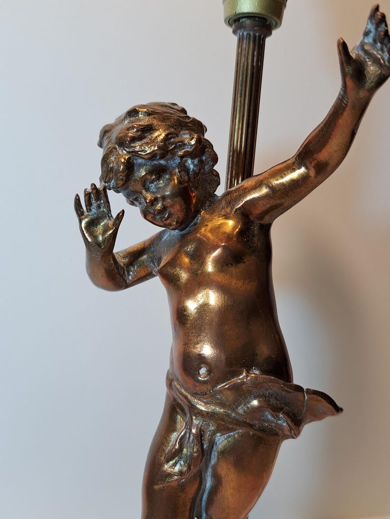 May include: A bronze-toned metal lamp base featuring a sculpted figure of a cherub with curly hair, arms raised in a dynamic pose, and a draped cloth around the waist. The figure is attached to a vertical fluted metal rod with a light socket.