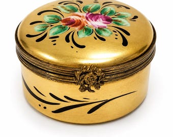 May include: A round gold-colored metal box with a hinged lid featuring a hand-painted floral design in pink, orange, and green. The box has black decorative brushstroke accents on the lid and sides, and a small ornate metal floral clasp.