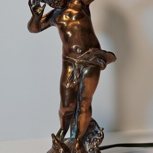 May include: A bronze-toned table lamp featuring a sculpted figure of a child with arms raised, standing on a circular base. The figure is draped in fabric around the waist and positioned in front of a vertical metal rod. The finish has metallic highlights.