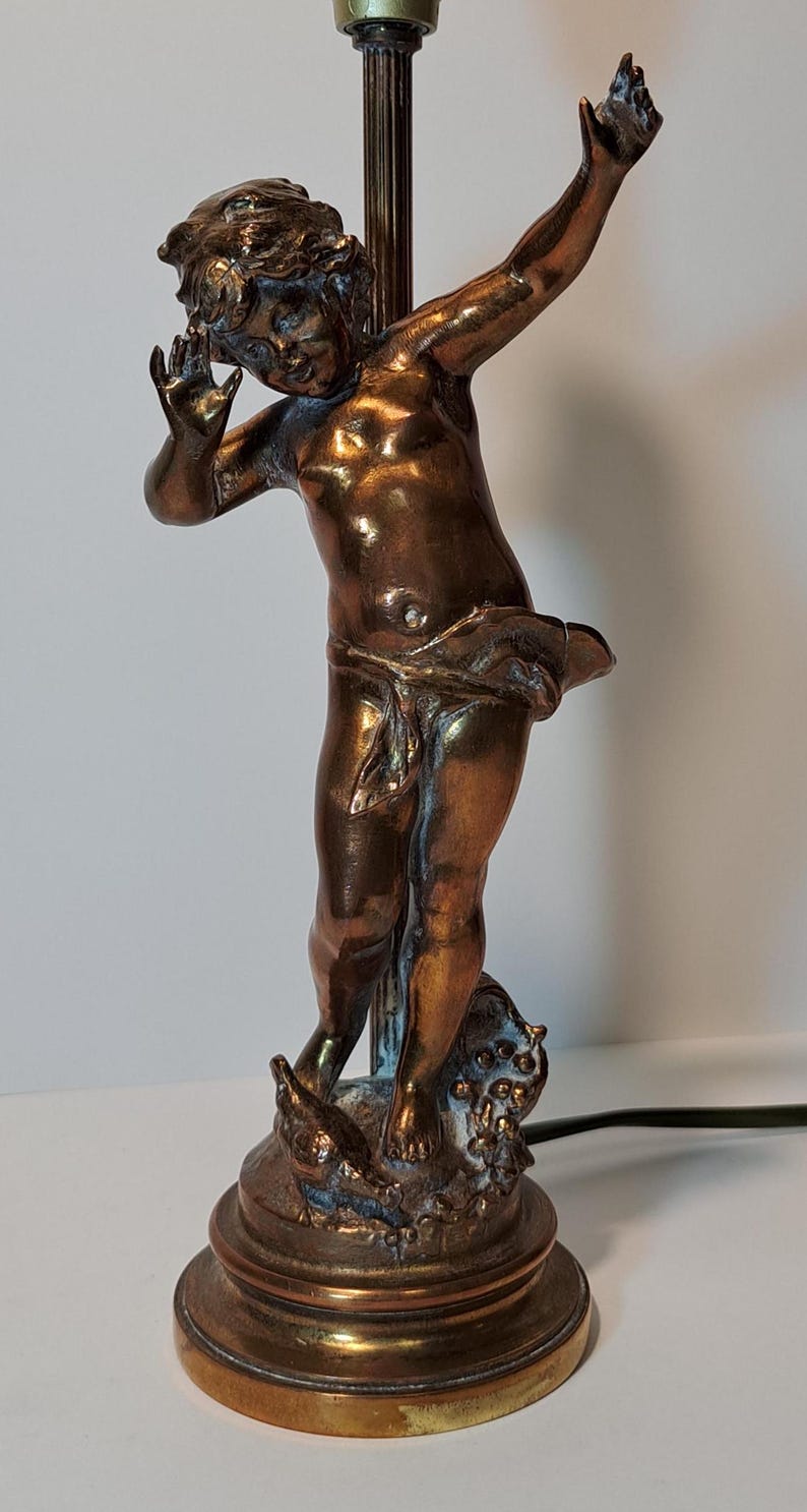 May include: A bronze-toned table lamp featuring a sculpted figure of a child with arms raised, standing on a circular base. The figure is draped in fabric around the waist and positioned in front of a vertical metal rod. The finish has metallic highlights.