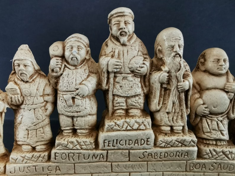 以下が含まれることがあります： 7体の幸運の神様を描いた、小さな白い陶器の置物のセットです。それぞれの置物は、ポルトガル語の言葉が刻まれたレンガの台座の上に立っています。言葉は「Justi&ccedil;a」、「Fortuna」、「Felicidade」、「Sabedoria」、「Boa Sa&uacute;de」です。
