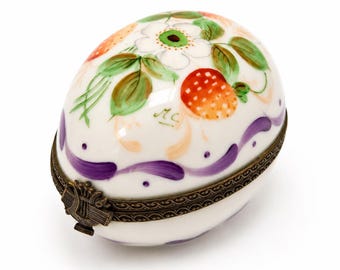 May include: An egg-shaped porcelain trinket box with a white base, hand-painted with green leaves, a white flower, and orange strawberries. It features a purple wavy line around the center and a decorative bronze-toned metal hinge with a lyre clasp.