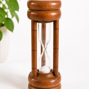 Può includere: Una clessidra in legno con finitura marrone lucida, dotata di una struttura cilindrica e due tappi arrotondati. Il bulbo centrale in vetro contiene sabbia bianca che scorre dalla camera superiore verso quella inferiore.