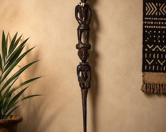 May include: A tall, dark brown wooden staff featuring two vertically stacked carved human figures. The top figure wears a light-colored beaded necklace, while the bottom figure is adorned with a blue beaded necklace. The staff has a smooth, tapered base.