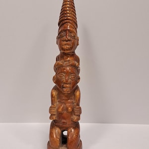 May include: A wooden sculpture of three figures stacked on top of each other. The top figure is wearing a conical hat and the bottom figure is kneeling. The sculpture is carved in a brown wood.