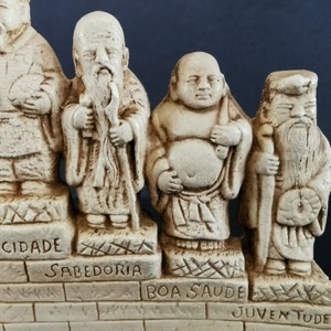 以下が含まれることがあります： 日本の神話を描いた、白い陶器製の七福神の置物セットです。それぞれの置物は、ポルトガル語が刻まれたレンガの台座の上に立っています。言葉は「Felicidade」、「Sabedoria」、「Boa Saude」、「Juventude」、「Longa Vida」、「Prosperidade」、「Abundancia」です。