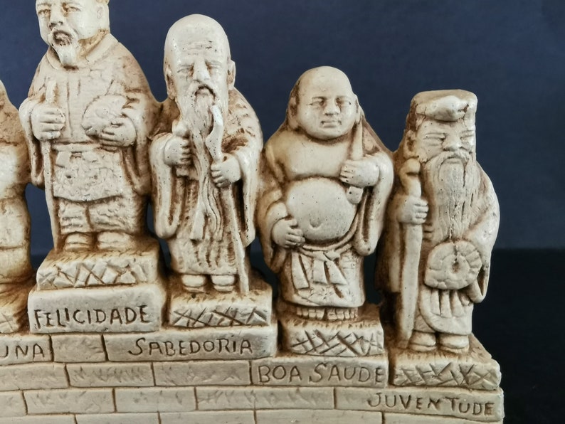 以下が含まれることがあります： 日本の神話を描いた、白い陶器製の七福神の置物セットです。それぞれの置物は、ポルトガル語が刻まれたレンガの台座の上に立っています。言葉は「Felicidade」、「Sabedoria」、「Boa Saude」、「Juventude」、「Longa Vida」、「Prosperidade」、「Abundancia」です。