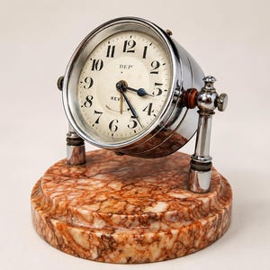 May include: A round analogue clock with a chrome finish and a white face featuring black numerals, the text DEP and REVEIL, and dark hands. The clock is mounted on two chrome pillars attached to a circular base made of polished brown marble.