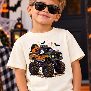 May include: A cream-colored short-sleeve t-shirt featuring a graphic of a monster truck decorated with Halloween elements, including a pumpkin, a ghost, bats, and a checkered pattern. The truck is orange, purple, and black with large tires.