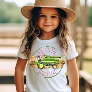 K&ouml;nnte beinhalten: Wei&szlig;es T-Shirt mit einem gr&uuml;nen und gelben M&auml;hdrescher-Motiv und dem Text "Dibs on the Buddy Seat". Das Shirt wird mit einer wei&szlig;en Hose und einem Strohhut getragen.