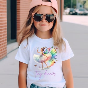 May include: A white t-shirt with a colorful illustration of a ballerina with butterfly wings. The text "tiny dancer" is printed in purple below the illustration.
