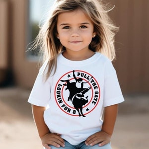 May include: A young girl wearing a white t-shirt with a black and red graphic that says "Lookin' for 8 Pull the Gate" and blue jeans. She is sitting on a wooden post.