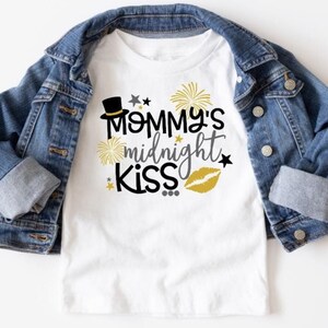May include: White t-shirt with a black and gold design that says "Mommy's midnight kiss" with a black top hat, gold stars, and a gold lip print.