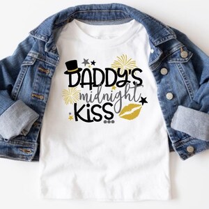 May include: White t-shirt with a black and gold design that says "Daddy's midnight kiss" with a top hat, fireworks, and a gold lip print.