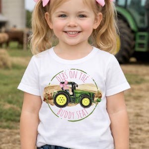 K&ouml;nnte beinhalten: Ein junges Kind tr&auml;gt ein wei&szlig;es T-Shirt mit einem gr&uuml;nen Traktorgrafik und dem Text "Dibs on the Buddy Seat". Das Kind l&auml;chelt und tr&auml;gt rosa Schleifen im Haar. Ein gr&uuml;ner Traktor ist im Hintergrund.