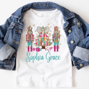 May include: White t-shirt with a colorful Christmas tree and two nutcracker figures. The text "Sophia Grace" is printed below the design.