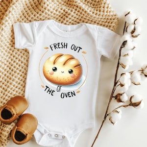 May include: White baby onesie with short sleeves featuring a cartoon bread with a smiling face and the text "Fresh out of the oven".