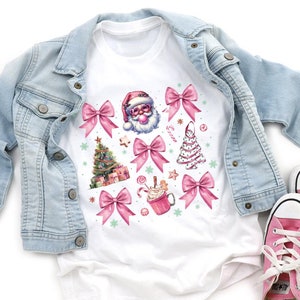 May include: White t-shirt with a pink and white Christmas design featuring Santa Claus, a Christmas tree, candy canes, bows, and a cup of hot chocolate.