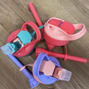 May include: Several colorful water bottle lids with attached straws. The lids are in shades of pink, blue, and teal. The straws are a solid pink color. The lids have a flip-top design and are made of plastic.