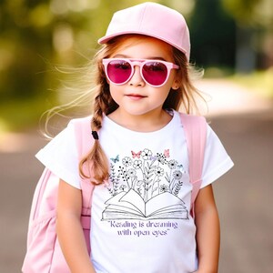 May include: A white t-shirt with a black and white illustration of an open book with flowers and butterflies. The text "Reading is dreaming with open eyes" is printed on the shirt.