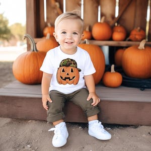May include: A child wearing a white t-shirt with a pumpkin graphic and a camouflage hat. The child is sitting in front of several orange pumpkins. The child is wearing olive green trousers and white trainers.