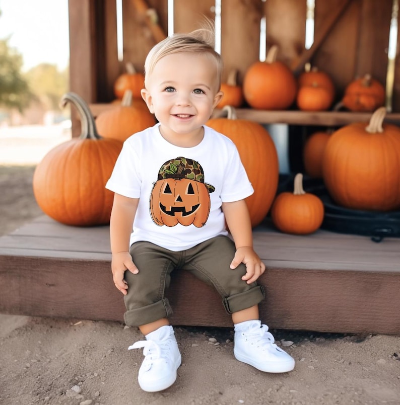 May include: A child wearing a white t-shirt with a pumpkin graphic and a camouflage hat. The child is sitting in front of several orange pumpkins. The child is wearing olive green trousers and white trainers.