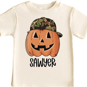 May include: Cream-coloured t-shirt with a smiling orange pumpkin wearing a camouflage baseball cap. The pumpkin has black eyes and a carved mouth. The name "SAWYER" is printed in black script below the pumpkin. A Halloween-themed graphic tee.