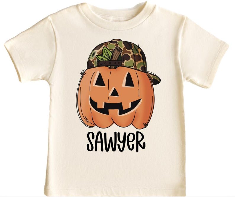 May include: Cream-coloured t-shirt with a smiling orange pumpkin wearing a camouflage baseball cap. The pumpkin has black eyes and a carved mouth. The name "SAWYER" is printed in black script below the pumpkin. A Halloween-themed graphic tee.