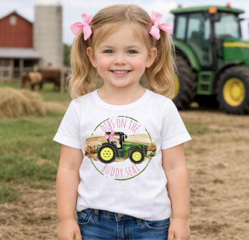 K&ouml;nnte beinhalten: Ein junges Kind tr&auml;gt ein wei&szlig;es T-Shirt mit einem gr&uuml;nen Traktorgrafik und dem Text "Dibs on the Buddy Seat". Das Kind l&auml;chelt und tr&auml;gt rosa Schleifen im Haar. Ein gr&uuml;ner Traktor ist im Hintergrund.