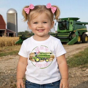 K&ouml;nnte beinhalten: Ein junges Kind tr&auml;gt ein wei&szlig;es T-Shirt mit einem gr&uuml;nen Traktorgrafik und dem Text "Dibs on the Buddy Seat". Das Kind hat blondes Haar, das zu Z&ouml;pfen mit rosa Schleifen gestylt ist. Im Hintergrund ist ein gr&uuml;ner M&auml;hdrescher zu sehen.