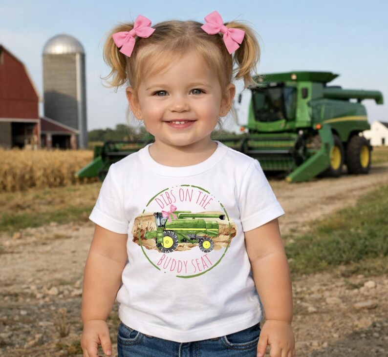 K&ouml;nnte beinhalten: Ein junges Kind tr&auml;gt ein wei&szlig;es T-Shirt mit einem gr&uuml;nen Traktorgrafik und dem Text "Dibs on the Buddy Seat". Das Kind hat blondes Haar, das zu Z&ouml;pfen mit rosa Schleifen gestylt ist. Im Hintergrund ist ein gr&uuml;ner M&auml;hdrescher zu sehen.