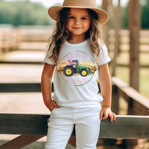 K&ouml;nnte beinhalten: Wei&szlig;es T-Shirt mit einem gr&uuml;nen und gelben Traktorgrafik und dem Text "Dibs on the Buddy Seat". Das Shirt wird von einer jungen Person getragen, die einen Strohhut und eine wei&szlig;e Hose tr&auml;gt.