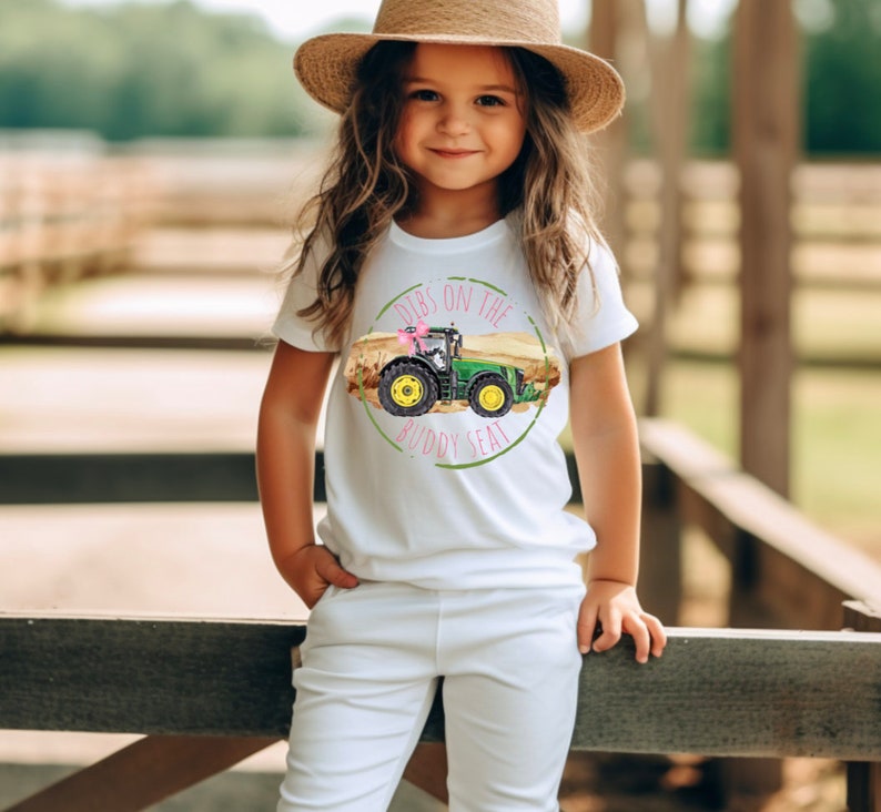 K&ouml;nnte beinhalten: Wei&szlig;es T-Shirt mit einem gr&uuml;nen und gelben Traktorgrafik und dem Text "Dibs on the Buddy Seat". Das Shirt wird von einer jungen Person getragen, die einen Strohhut und eine wei&szlig;e Hose tr&auml;gt.
