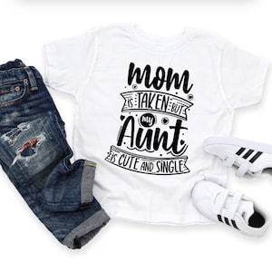 May include: A white t-shirt with the text "Mom is taken but my Aunt is cute and single" in black script. The shirt is paired with blue denim jeans and white sneakers with black stripes.