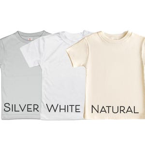 Puede incluir: Tres camisetas de manga corta en diferentes colores: plata, blanco y natural. Las camisetas est&aacute;n dispuestas en plano y la parte inferior de cada camiseta tiene el nombre del color impreso en negro.