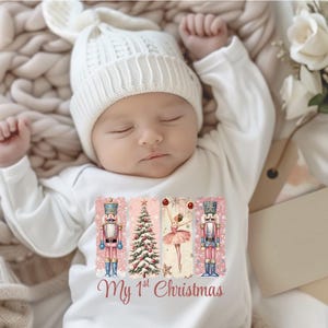 May include: A sleeping baby wearing a white knit cap and a long-sleeved onesie. The onesie features a festive design with a nutcracker, Christmas tree, and ballerina, with the text "My 1st Christmas" in a script font.