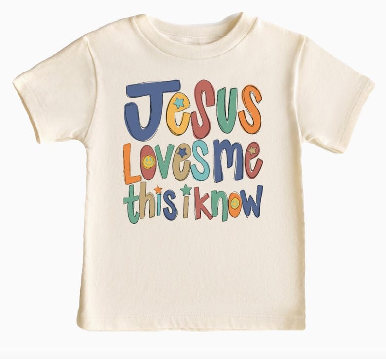 Puede incluir: Una camiseta blanca con un gr&aacute;fico colorido que dice "Jes&uacute;s me ama, esto lo s&eacute;". El gr&aacute;fico incluye estrellas y caras sonrientes.