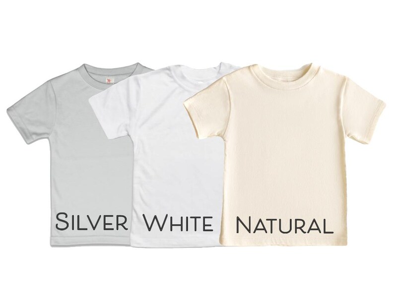 May include: Three short-sleeved t-shirts in silver, white, and natural colours. The word "SILVER" is printed in black below the silver shirt, "WHITE" below the white shirt, and "NATURAL" below the natural shirt. The shirts are plain and have a crew neck.