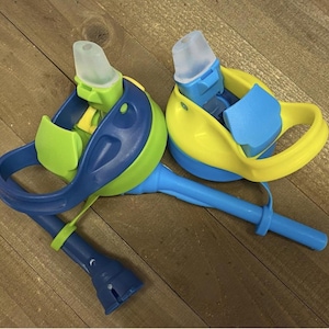 May include: Two colorful water bottles with blue, green, and yellow accents. Each bottle has a clear, soft spout and a handle. The bottles are designed for easy drinking and are likely for children.
