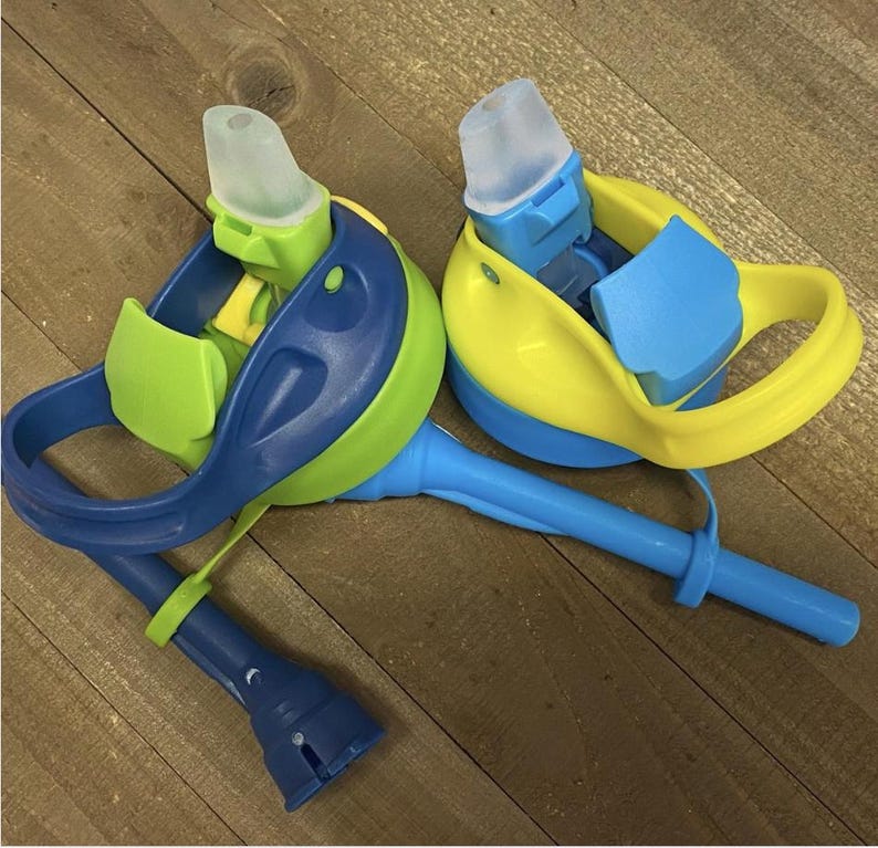 May include: Two colorful water bottles with blue, green, and yellow accents. Each bottle has a clear, soft spout and a handle. The bottles are designed for easy drinking and are likely for children.