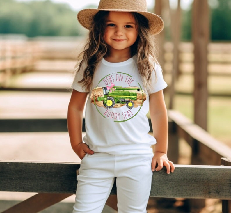 K&ouml;nnte beinhalten: Wei&szlig;es T-Shirt mit einem gr&uuml;nen und gelben M&auml;hdrescher-Motiv und dem Text "Dibs on the Buddy Seat". Das Shirt wird mit einer wei&szlig;en Hose und einem Strohhut getragen.