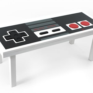 Op de afbeelding: Een witte salontafel in de vorm van een klassieke NES-controller. De tafel heeft een zwart blad met een grijze streep en rode knoppen. De tafel heeft vier witte poten.