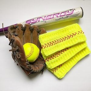 May include: Three yellow crocheted headbands with red stitching, resembling baseball stitches, are stacked on top of each other. A brown leather softball glove with a yellow softball is in the background. A purple and white softball bat is also in the background.
