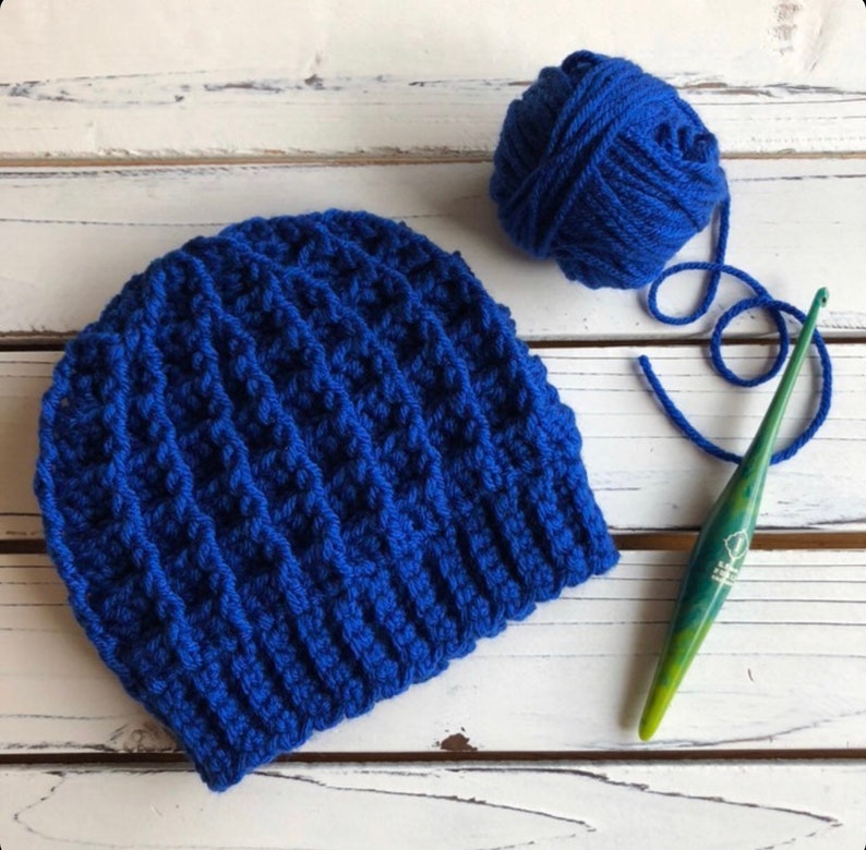 May include: A blue crocheted beanie hat with a ball of blue yarn and a green crochet hook.