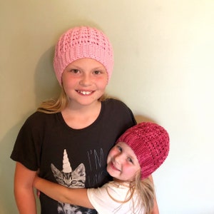 May include: Two girls wearing crocheted hats. The girl in the front is wearing a pink hat and the girl in the back is wearing a red hat. The girl in the back is wearing a black t-shirt with a unicorn cat graphic and the text "I'm Unique".