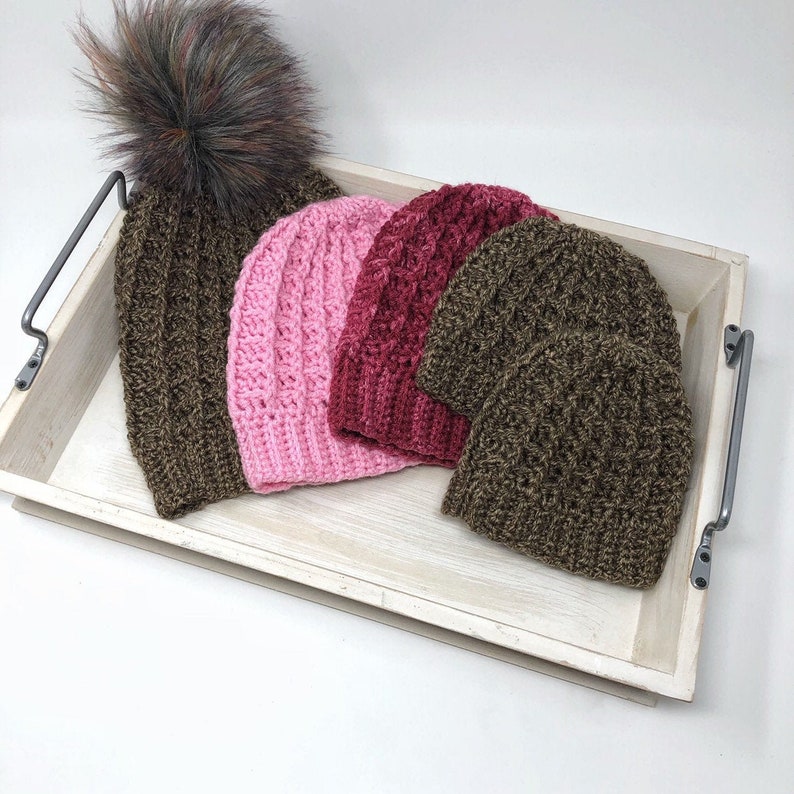 May include: Three crocheted hats in a wooden tray. One hat is brown, one is pink, and one is burgundy. The brown hat has a fluffy pompom on top.