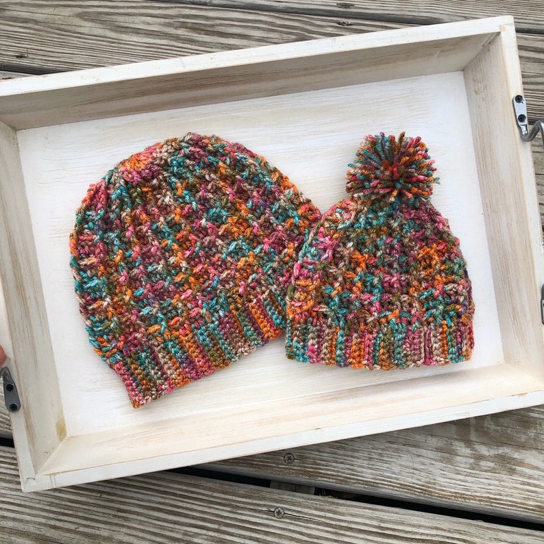 May include: Two crocheted hats in a variety of fall colors, including orange, brown, pink, and teal. The hats are sitting in a wooden tray.