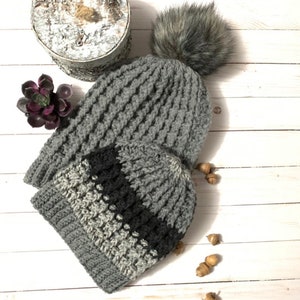 May include: Two gray crocheted hats with a black stripe on the smaller hat. The larger hat has a faux fur pom pom on top. The hats are on a white wooden surface with acorns scattered around.